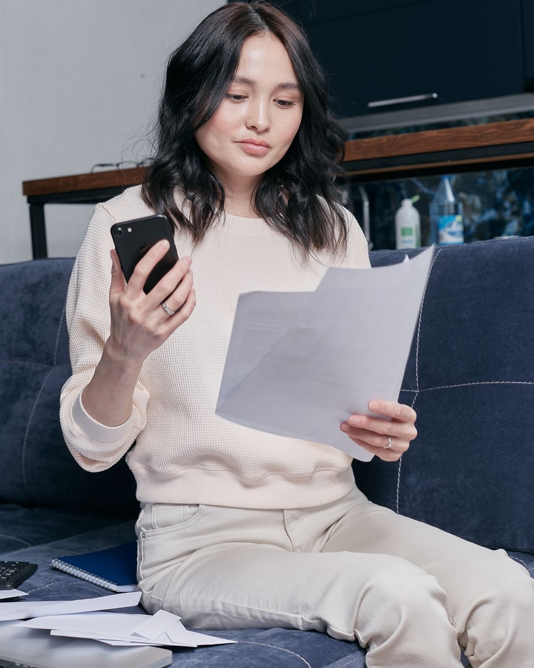 A young woman sits alone on her couch, phone in one hand and a paper statement in the other, sorting out her own finances without an advisor.